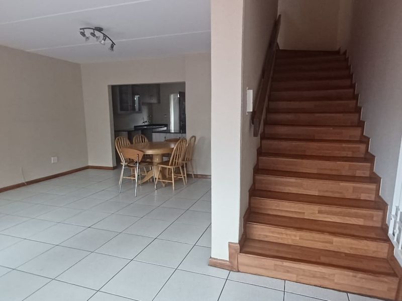Open plan living area featuring tiled flooring and a wooden staircase with a security gate, leading towards a dining space and kitchen.
