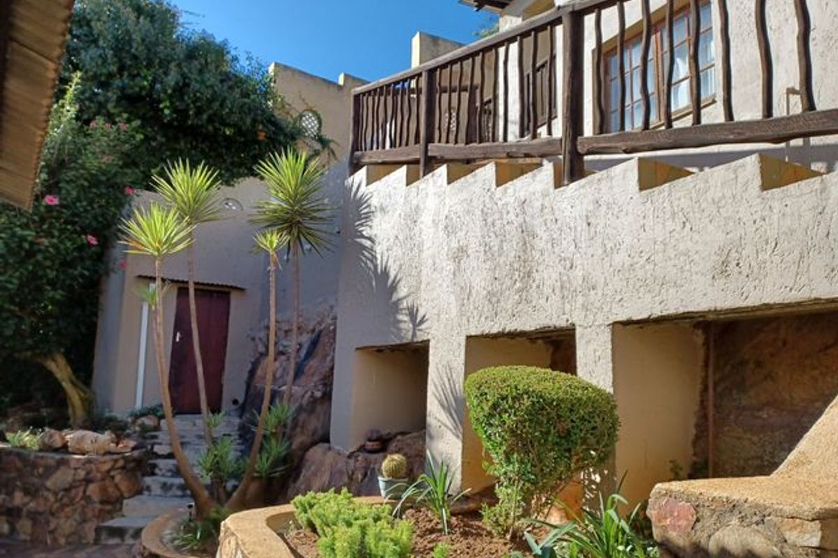 Rear exterior view featuring a multi-level garden with stone retaining walls, a paved courtyard, and a timber balcony overlooking the landscaped area.