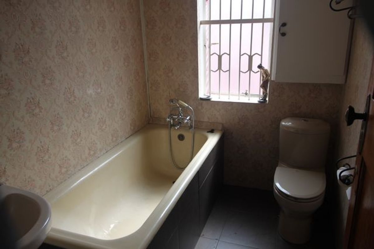 A dated bathroom featuring a bathtub, toilet, and basin, with patterned wallpaper and security bars on the window.