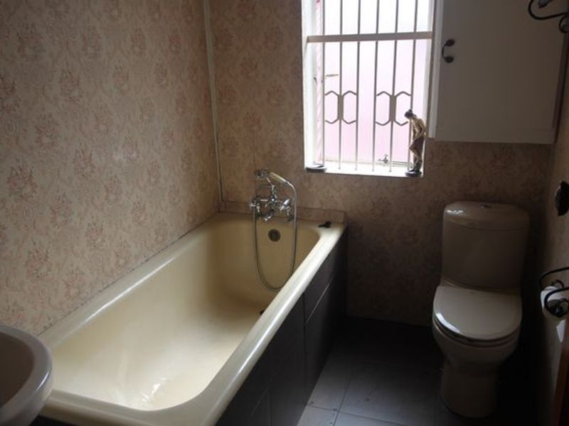 A dated bathroom featuring a bathtub, toilet, and basin, with patterned wallpaper and security bars on the window.