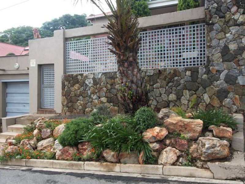 Front exterior view featuring a stone-clad boundary wall, integrated garage, and a landscaped rock garden with a central palm tree.