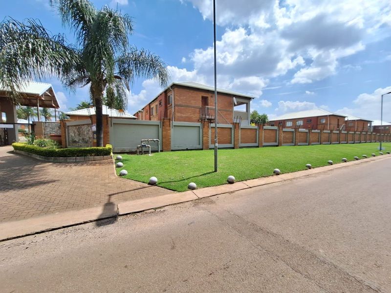 Street view of the apartment complex featuring a secure brick perimeter wall with electric fencing, well-maintained lawn, and paved driveway access.