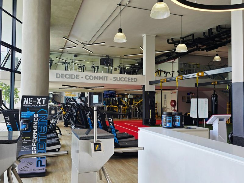 Spacious modern gym facility featuring a reception area, turnstile access control, mezzanine level, and large windows providing ample natural light.
