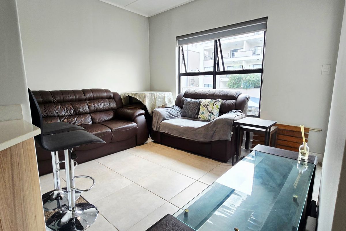 A functional living room featuring neutral tiled flooring and a large window with a roller blind, providing natural light to the space.