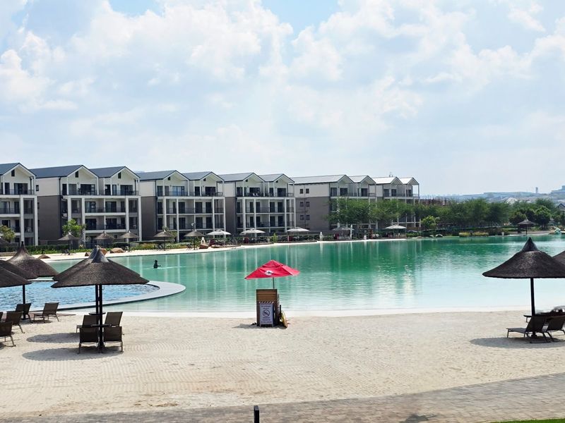 A large communal swimming pool with a sandy beach area, surrounded by modern apartment buildings.