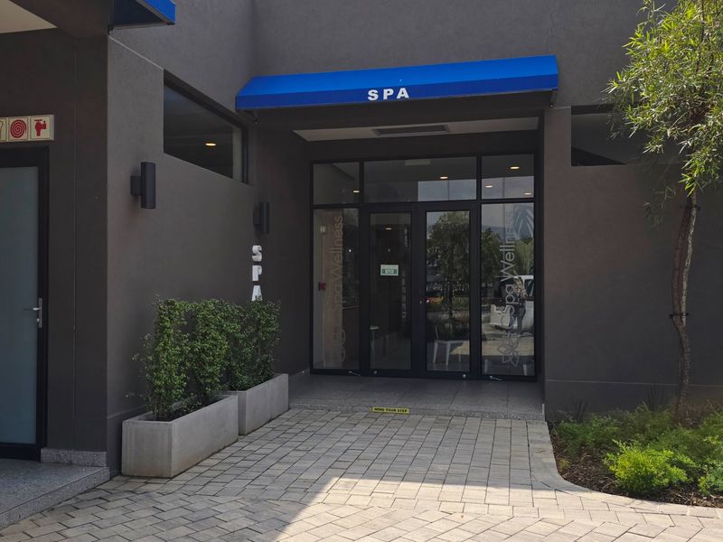 The entrance to the spa facility features a modern glass door, a blue awning, and a paved walkway with integrated concrete planters.