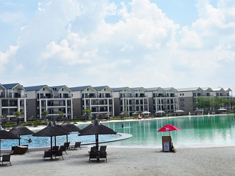 A view of a large communal swimming pool and sandy beach area set against a modern apartment complex.