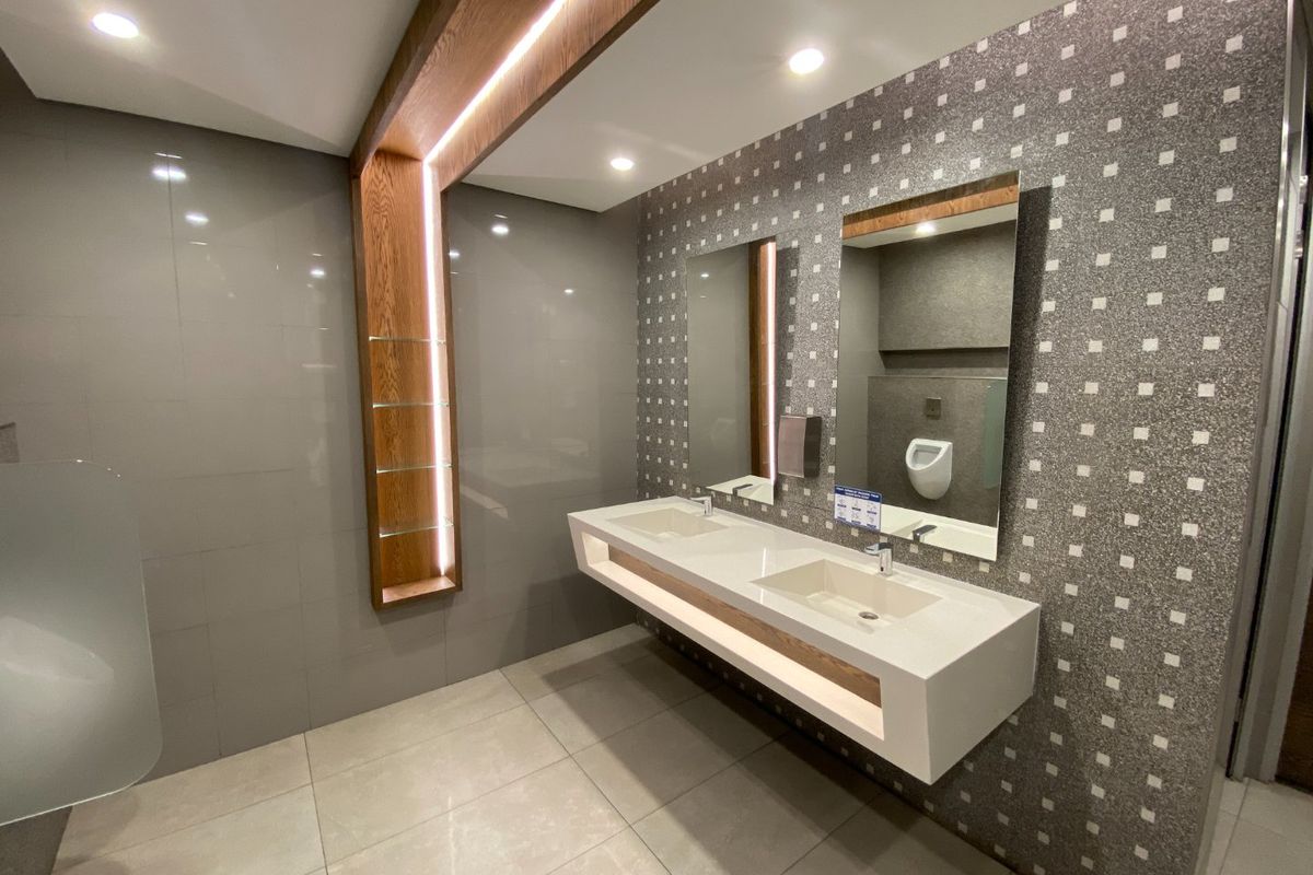 Modern commercial bathroom featuring a sleek wall-mounted double vanity, integrated sinks, contemporary tiled finishes, and recessed LED accent lighting.