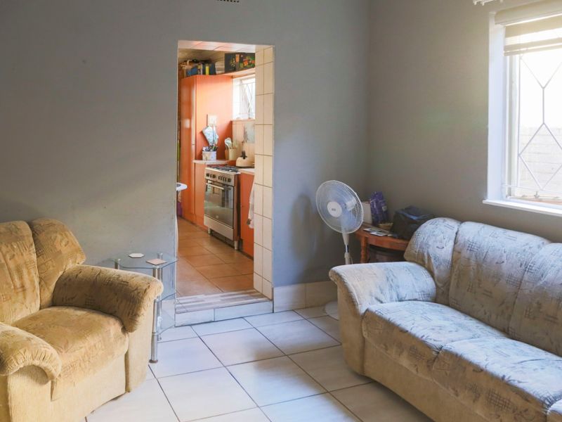 A living room area with tiled flooring and a direct view into the adjacent kitchen, featuring neutral wall tones and natural light.