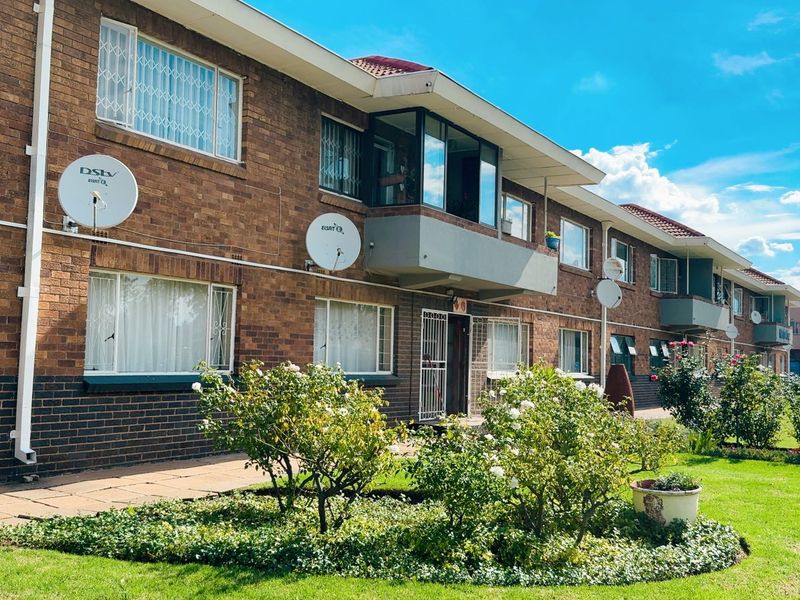 A view of the front facade of a brick apartment building featuring a landscaped garden, paved walkway, and balconies.