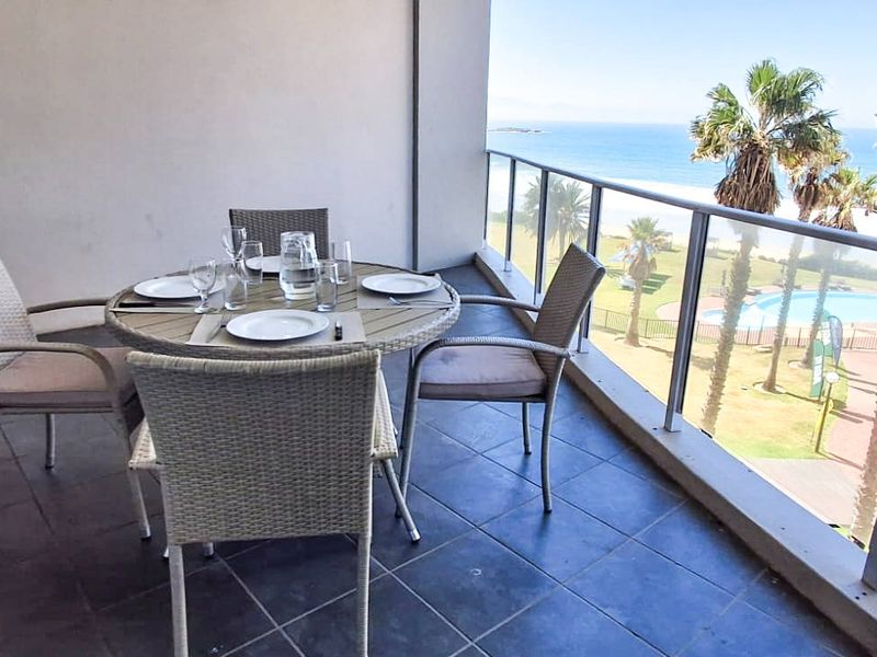 A spacious balcony featuring a glass balustrade and tiled flooring, offering scenic coastal and pool area views.