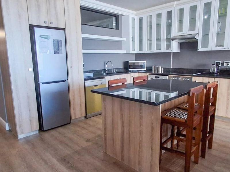 Modern open-plan kitchen featuring a central island, granite countertops, light wood cabinetry, and integrated appliances, seamlessly flowing into the adjacent hallway.