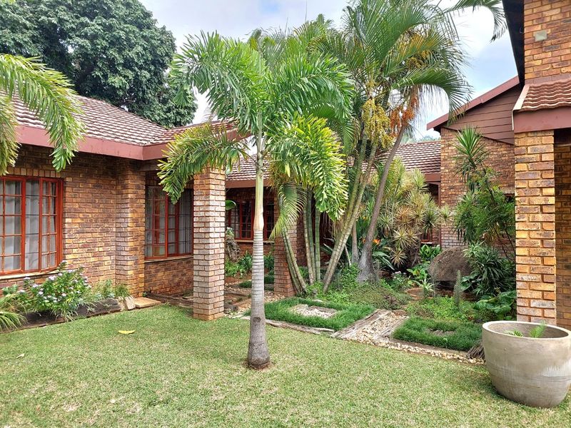 A well-maintained rear garden area featuring face brick exterior walls, tiled roofing, and established landscaping with mature palm trees and brick pathways.