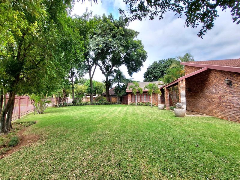 A well-maintained rear garden featuring a spacious lawn, mature trees, and a brick-built house with a covered patio area.