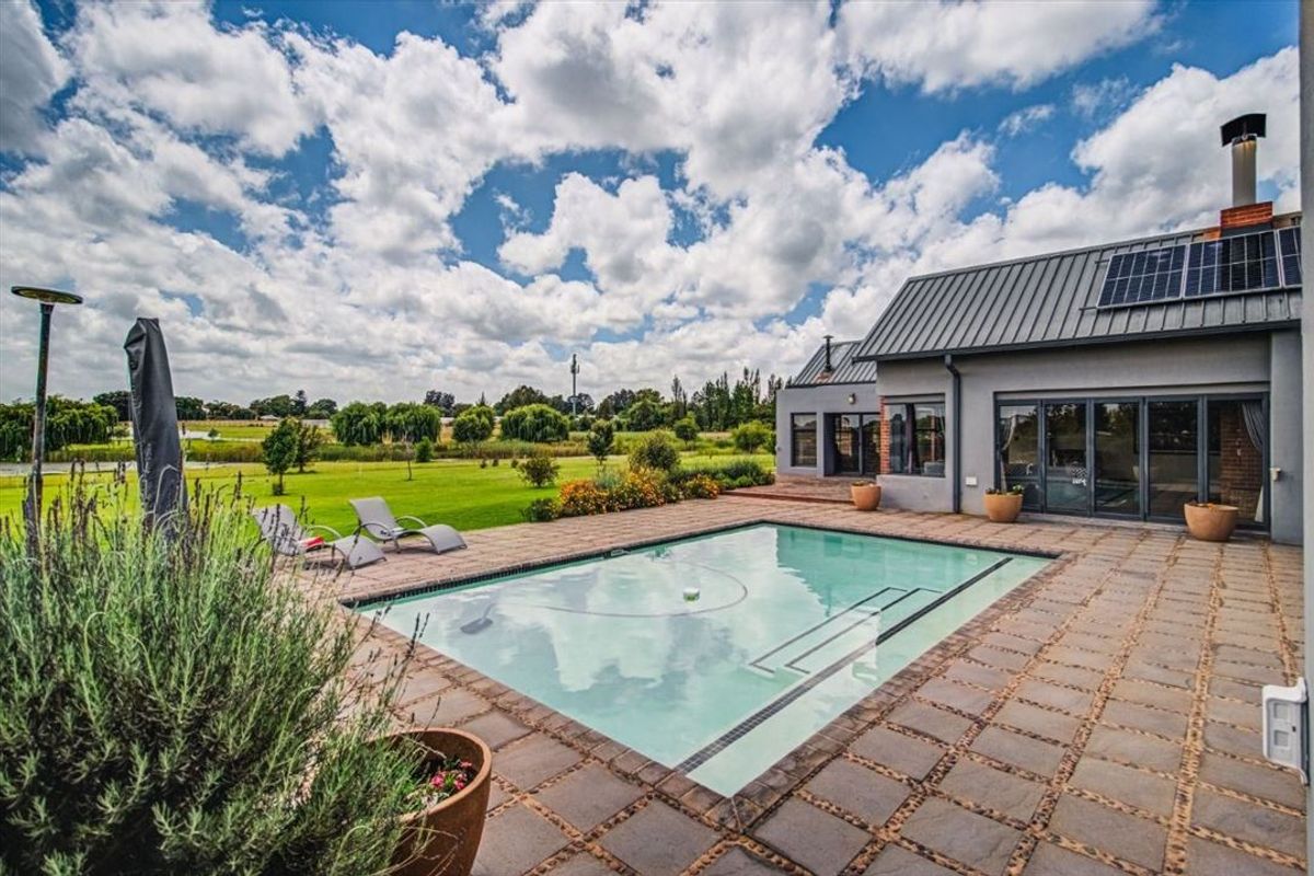 A well-maintained outdoor pool area featuring a paved patio, modern metal roofing with solar panels, and expansive sliding glass doors leading to the home.