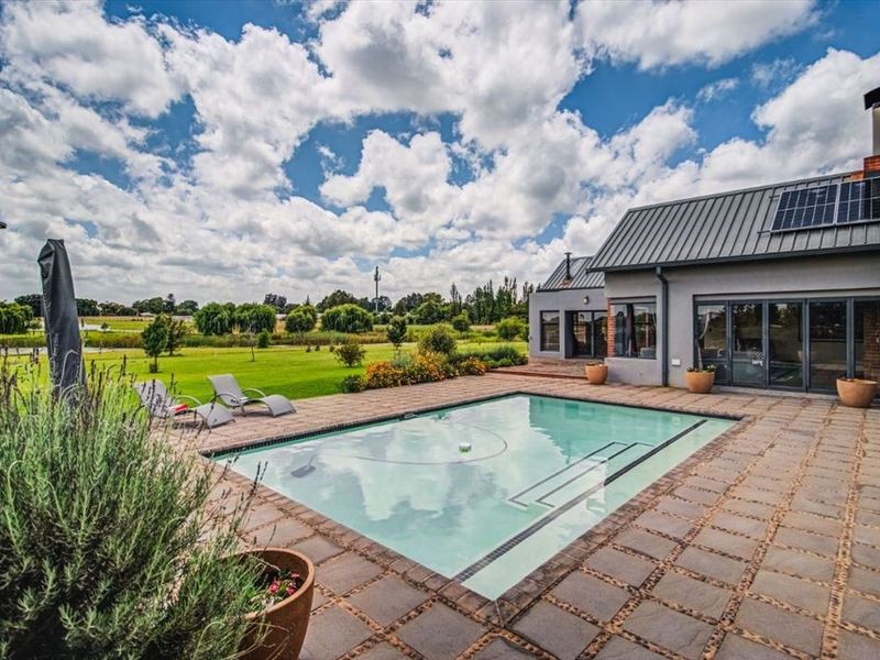 A well-maintained outdoor pool area featuring a paved patio, modern metal roofing with solar panels, and expansive sliding glass doors leading to the home.