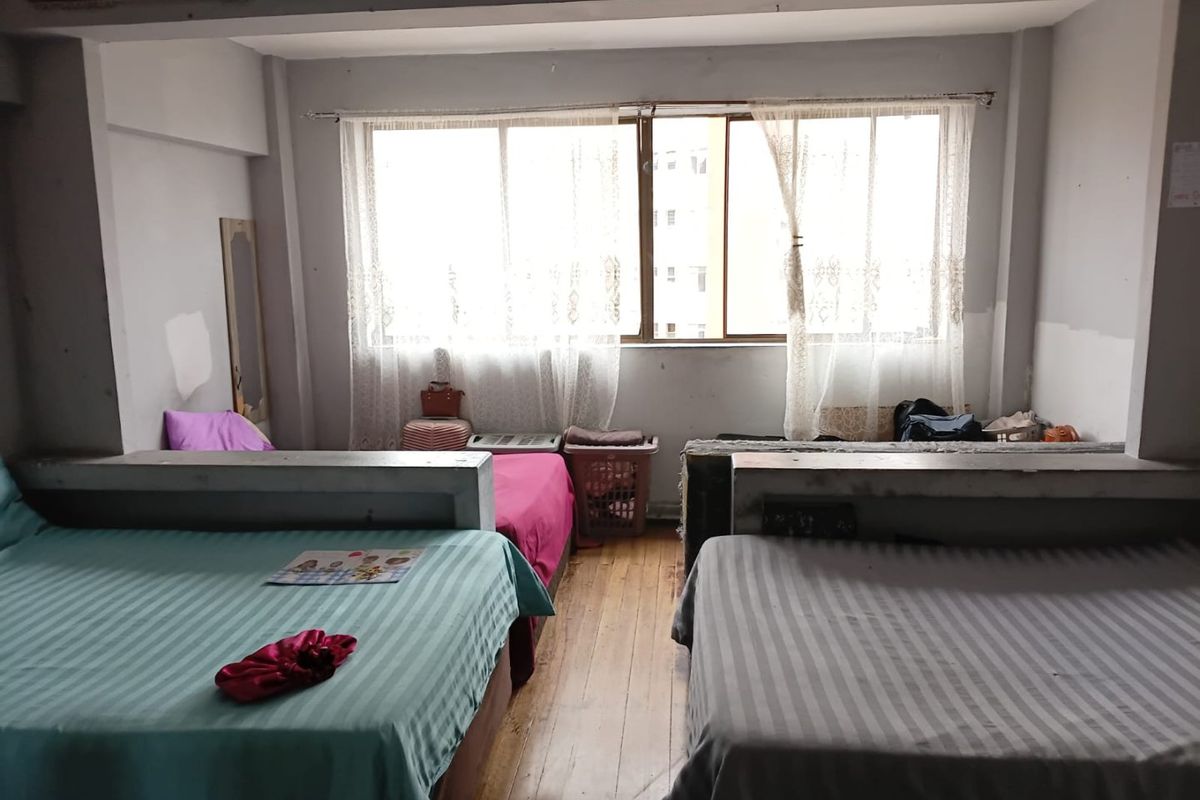 A bedroom featuring hardwood flooring and large windows, currently configured with multiple beds. The space requires cosmetic updates and wall repairs.