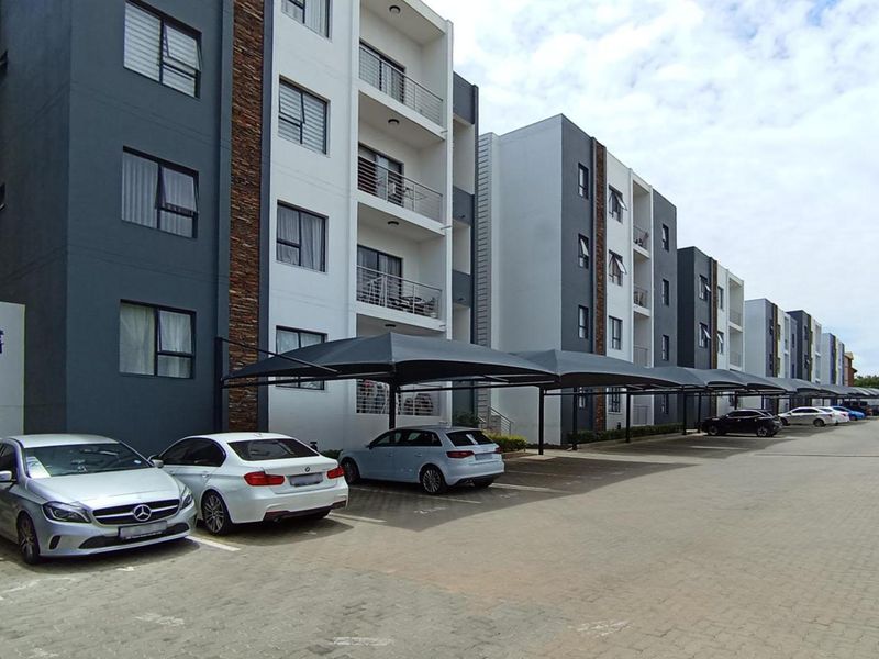 Exterior view of a modern multi-story apartment complex featuring paved parking bays with shade cloth carports and private balconies.
