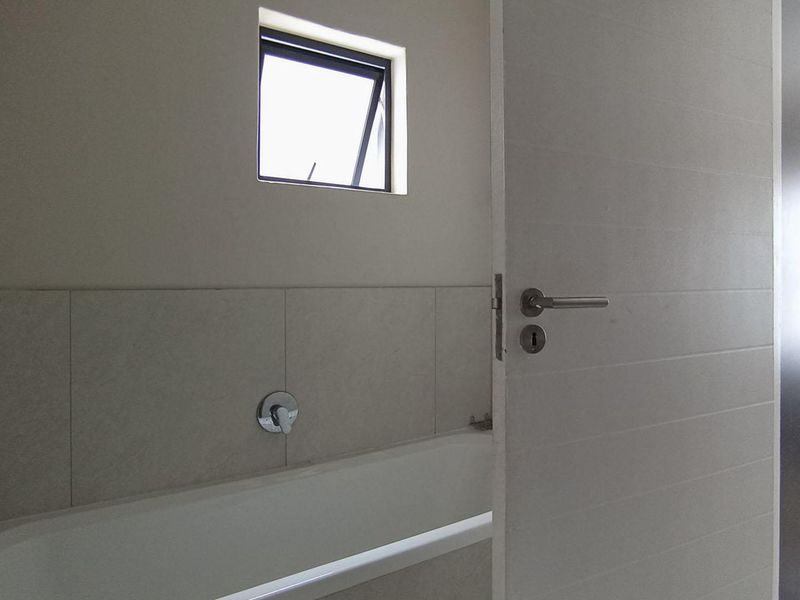 A clean, neutral-toned bathroom featuring a built-in bathtub, tiled wall finishes, and a top-hung window, with a view into the adjacent bedroom.
