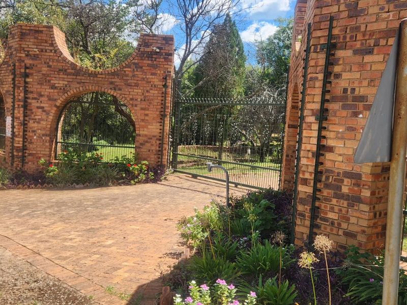 Entrance to the farm property featuring decorative brick gate pillars, a secure metal gate, and a paved driveway surrounded by established garden landscaping.