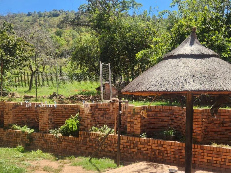 A landscaped garden area featuring a traditional thatched lapa, a curved brick retaining wall, and perimeter wire fencing set against a natural hillside backdrop.