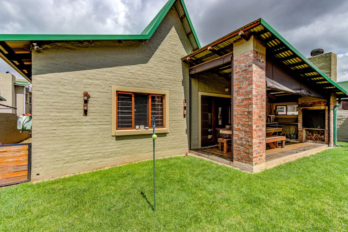 Rear view of the property featuring a covered patio with a built-in braai, exposed brick pillar, and a well-maintained lawn.