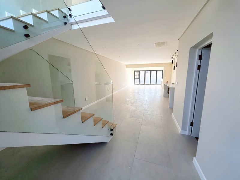 Spacious open-plan living area featuring a modern glass balustrade staircase, large format floor tiles, and sliding doors leading to the exterior.