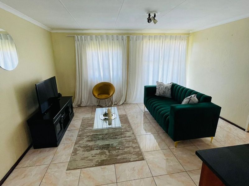 A living room area featuring tiled flooring, neutral wall finishes, and large windows with curtain tracks, providing a functional and well-lit space.