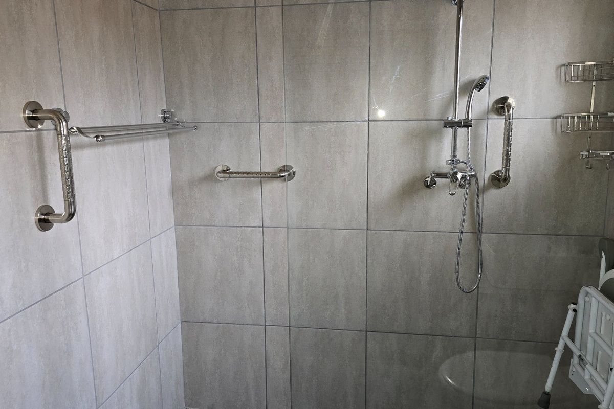 A modern tiled shower enclosure featuring a wall-mounted shower head, chrome mixer tap, and multiple safety grab rails for accessibility.