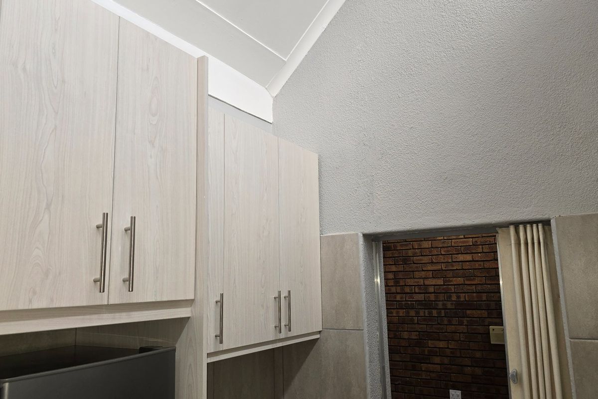 A functional kitchen area featuring light wood-grain cabinetry, high vaulted ceilings, and tiled splashbacks, leading to an adjacent brick-walled space.