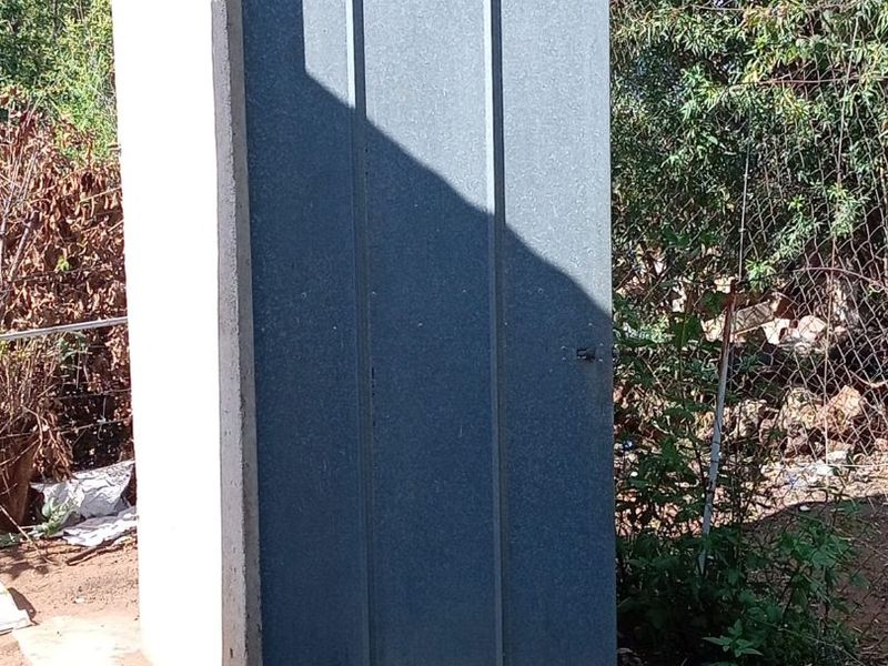 A standalone outdoor utility structure featuring a corrugated metal door and concrete base, situated in a garden setting.