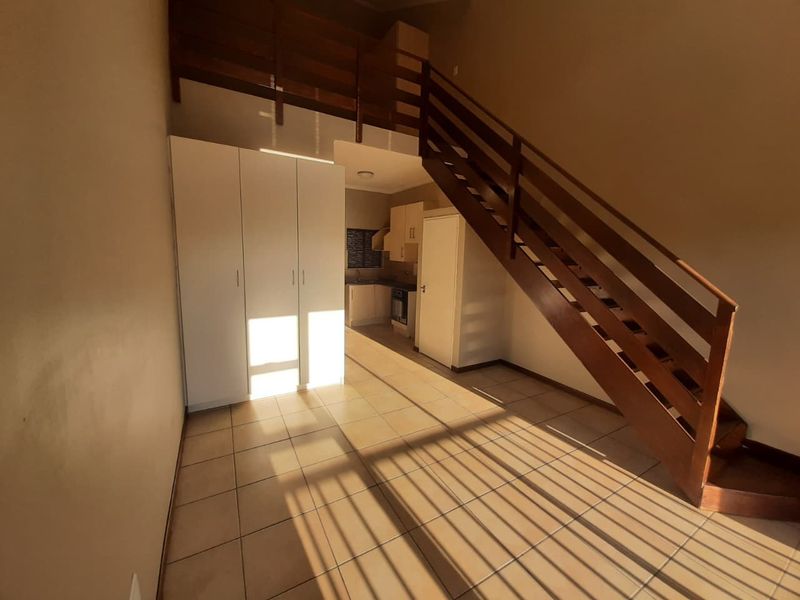 Open-plan living area featuring a wooden staircase leading to a mezzanine level, tiled flooring, and a compact kitchen with built-in cabinetry.