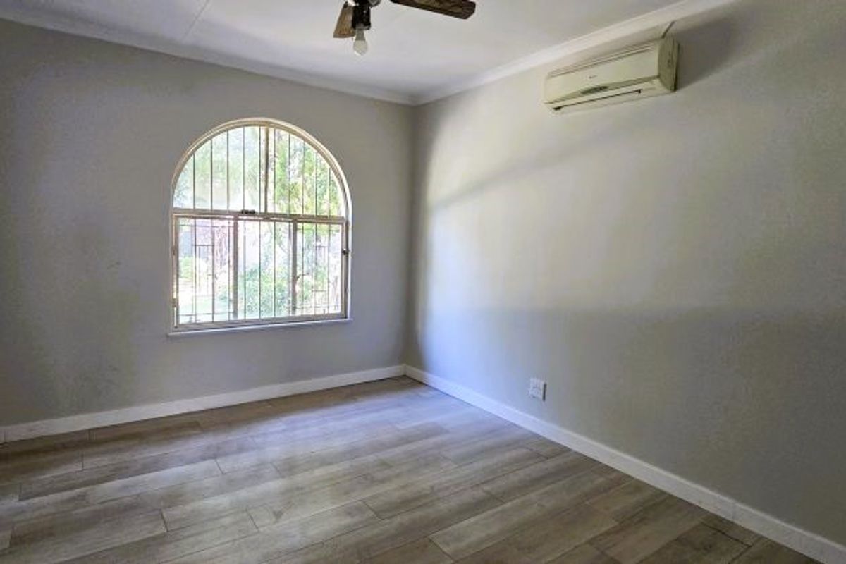An empty bedroom featuring laminate flooring, an arched window with security bars, a ceiling fan, and a wall-mounted air conditioning unit.