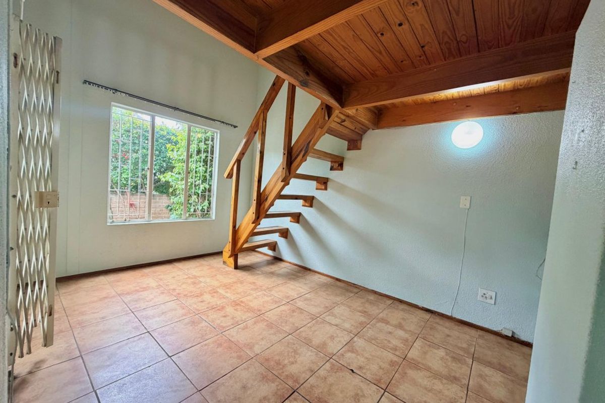 An open-plan living area featuring a wooden staircase leading to a mezzanine level, tiled flooring, and security features on the window and entrance.