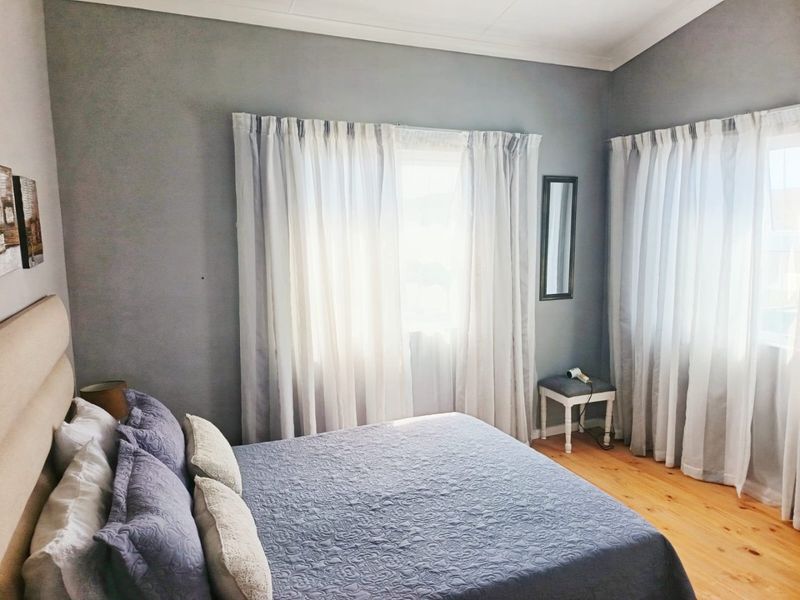 A well-lit bedroom featuring warm timber flooring, neutral grey walls, and large windows dressed with soft curtains.