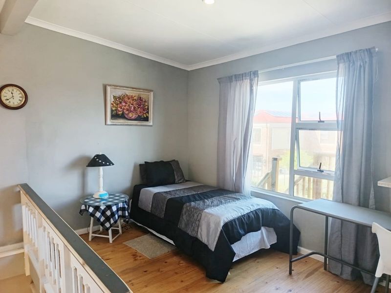A bedroom featuring timber flooring, neutral wall finishes, and a large window providing natural light, situated adjacent to an internal staircase balustrade.