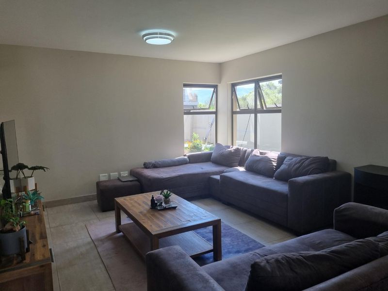 A comfortable living room featuring tiled flooring, neutral walls, and large aluminium-framed windows that provide natural light.