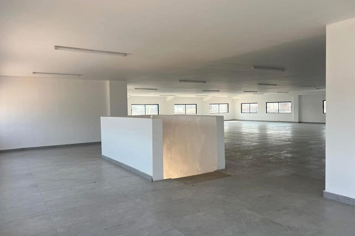 A large, open-plan commercial office space featuring neutral tiled flooring, ample natural light from multiple windows, and functional overhead strip lighting.