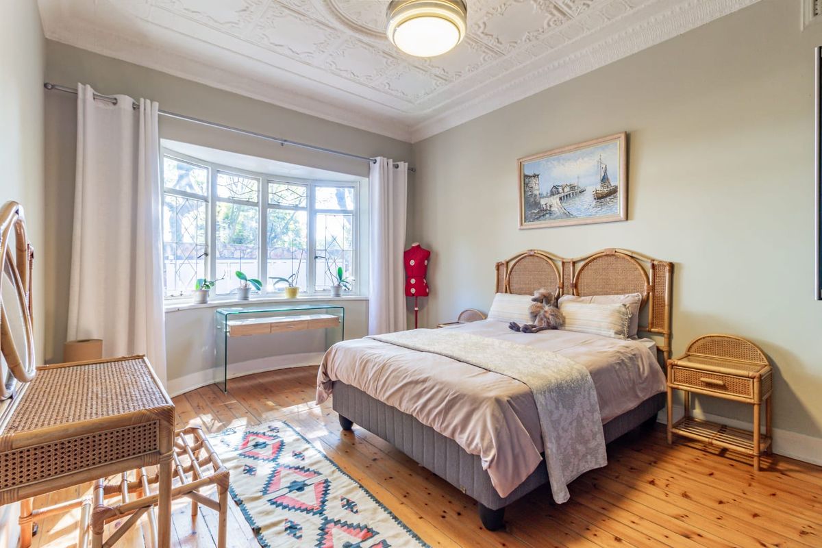 A well-maintained bedroom featuring original timber flooring, a decorative plaster ceiling with a central rose, and a large bay window providing natural light.