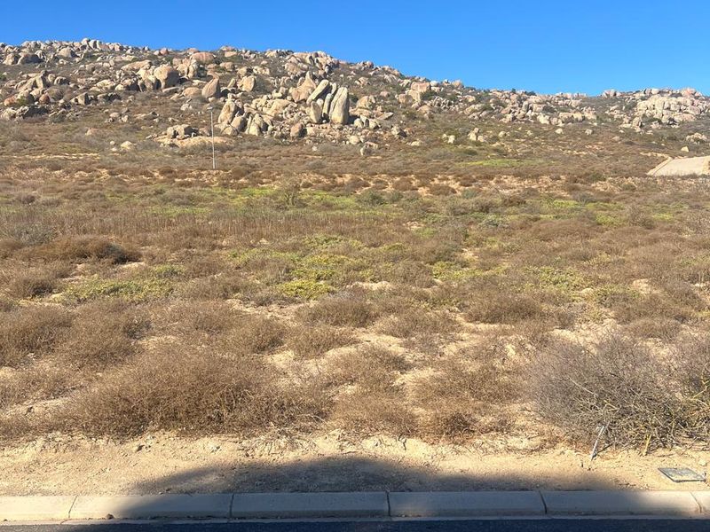 A vacant residential plot featuring natural rocky terrain and native vegetation, bordered by a paved road and concrete curb.