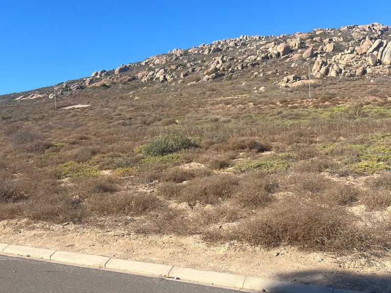 A view of the vacant residential land plot from the adjacent asphalt road, featuring natural rocky terrain and native vegetation.