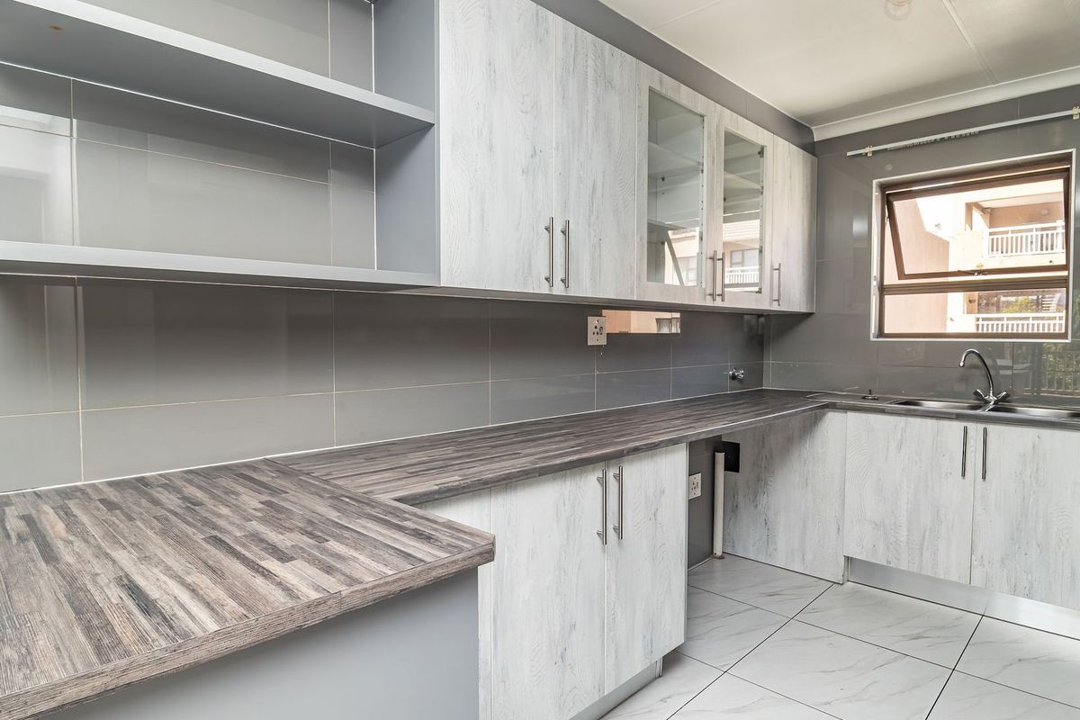 A modern, clean kitchen featuring light wood-grain cabinetry, dark laminate countertops, grey tiled splashbacks, and tiled flooring with an integrated sink.
