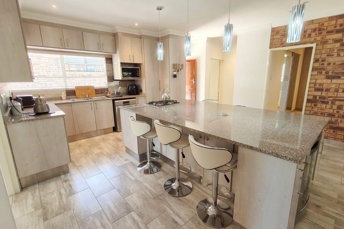 Modern kitchen featuring a large granite-topped island, light wood cabinetry, gas hob, and a stylish exposed brick feature wall with tiled flooring throughout.