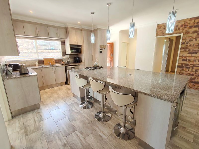 Modern kitchen featuring a large granite-topped island, light wood cabinetry, gas hob, and a stylish exposed brick feature wall with tiled flooring throughout.