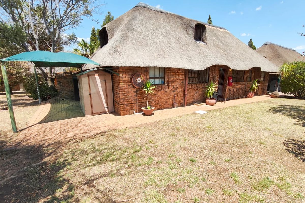 Rear view of a face brick house featuring a traditional thatch roof, security bars on windows, and a paved outdoor area with a shade sail.
