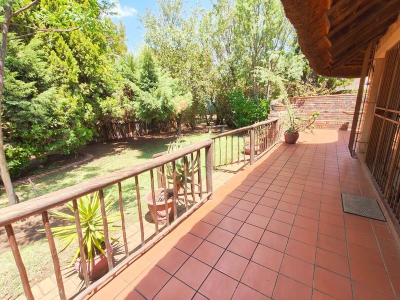 A covered patio featuring terracotta tiled flooring and a timber balustrade, overlooking a lush, established garden with mature trees.
