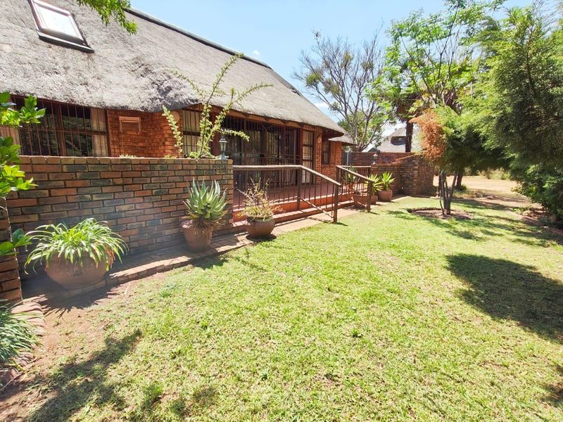 A rear view of a face brick house featuring a traditional thatch roof, a raised brick patio area, and a well-maintained garden.