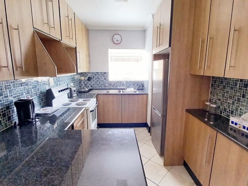 A functional galley-style kitchen featuring wood-look cabinetry, dark granite countertops, mosaic tile splashbacks, and tiled flooring.