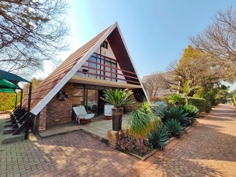 A charming A-frame house featuring a brick facade, a front patio, and a balcony, set within a well-maintained garden and paved driveway area.