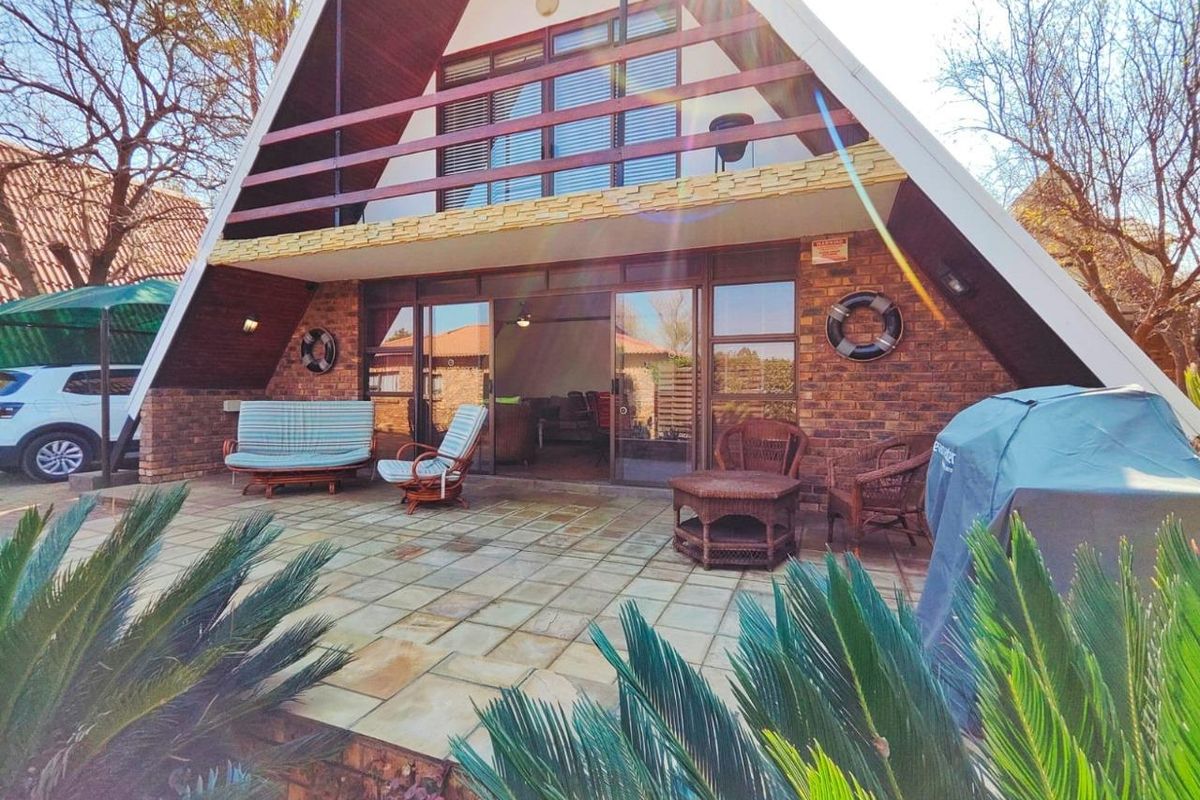 A charming A-frame house featuring a spacious stone-tiled rear patio, brick exterior, and an upper-level balcony overlooking the garden area.